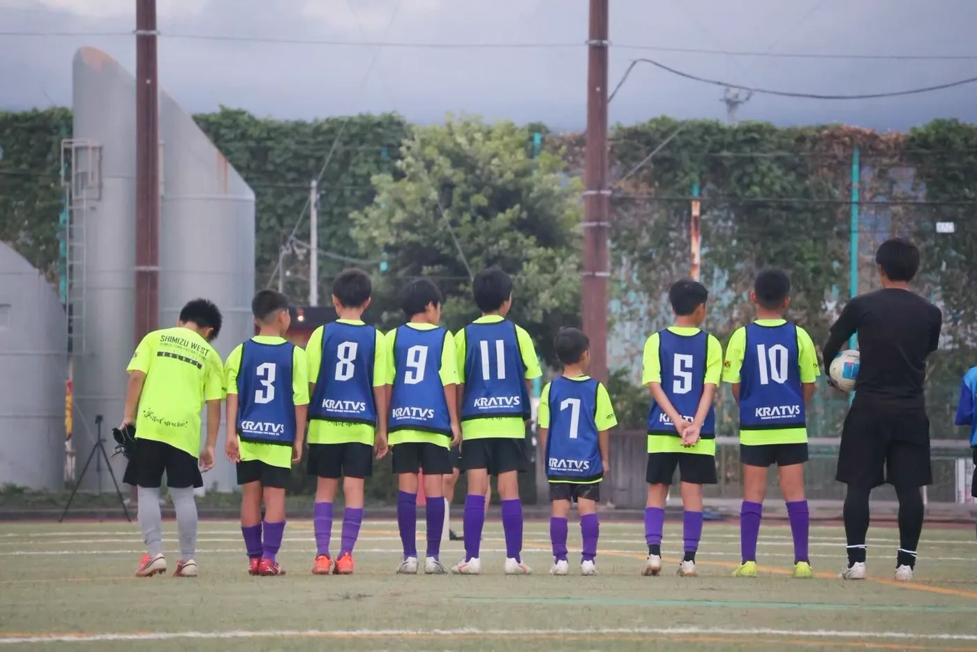 蛍光イエローの練習着の上に青いビブスを重ねて整列するサッカー少年団の子どもたちの後ろ姿
